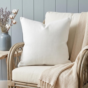 May include: A large, square white pillow with a textured surface sits on a cream-colored chair. A beige throw blanket with fringe is draped over the chair. A vase of lavender and cotton stems is on a side table.
