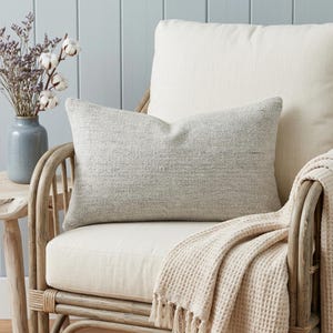 Peut inclure: Un oreiller rectangulaire gris clair avec un tissage texturé est posé sur une chaise crème. Une couverture crème avec des franges est drapée sur la chaise. Un vase avec des fleurs séchées est sur une table d'appoint en bois.