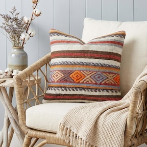 Puede incluir: Un cojín decorativo con un diseño tejido con rayas horizontales en tonos de rojo, naranja, marrón y gris. El cojín está colocado sobre una silla color crema con una estructura de ratán tejido y una manta color crema con flecos.