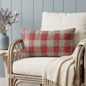 May include: A rectangular pillow with a red and gray plaid pattern rests on a cream-colored cushion. The pillow is on a wicker chair with a cream-colored blanket draped over the arm. A vase with lavender and cotton stems sits on a small wooden table.