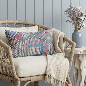 May include: A rectangular decorative pillow with a geometric pattern in shades of blue, pink, and gray rests on a cream-colored cushion. The pillow is on a wicker chair with a cream-colored blanket draped over it. A vase of flowers sits on a small wooden table.