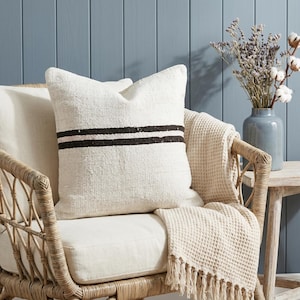May include: A white textured pillow with two dark brown stripes, resting on a wicker chair with a cream-colored cushion. A cream-colored waffle-weave throw with fringe is draped over the chair. A blue vase with dried flowers sits nearby.
