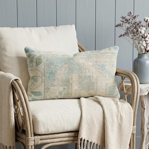 Peut inclure: Un coussin rectangulaire avec un motif bleu et beige délavé repose sur une chaise crème. Une couverture crème avec des franges est drapée sur la chaise. Un vase de fleurs séchées est posé sur une petite table d'appoint.