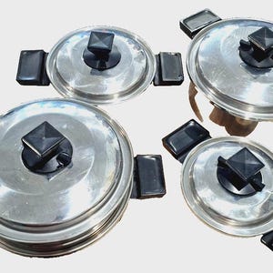 May include: A collection of stainless steel saucepans and lids. Each lid features a black square handle. The pans have black rectangular side handles. The set includes pans of different sizes, ideal for cooking.
