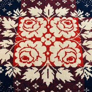 May include: Close-up of a textile with a repeating pattern. The design features red roses and white leaves on a cream background, framed by a dark blue and burgundy border with white snowflake and cross accents. The overall aesthetic is vintage and decorative.