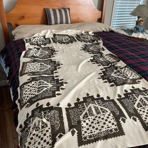 May include: A cream-colored textile with a repeating geometric pattern in dark brown. The pattern includes squares, rectangles, and other shapes. The textile is draped over a bed with a wooden headboard and a plaid blanket.