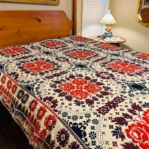 May include: A vintage-style bedspread with a floral pattern. The cream-colored blanket features large red roses, dark blue accents, and a decorative fringe. The bed frame is made of light-colored wood.
