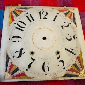 May include: Antique clock face with black numerals on a cream background. The clock face has decorative gold, red, and black accents in the corners. The center has a gold ring of dots. The clock face measures approximately 12 inches square.