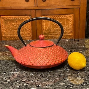 May include: A red cast iron teapot with a black handle and a textured surface. The teapot is sitting on a gray countertop next to a yellow lemon.