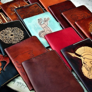 Könnte beinhalten: Eine Sammlung von ledergebundenen Tagebüchern mit verschiedenen Designs, darunter ein Drache, ein Pferd, ein Krieger und eine Karte. Die Tagebücher sind alle braun oder schwarz und haben ein rustikales, handgemachtes Aussehen.