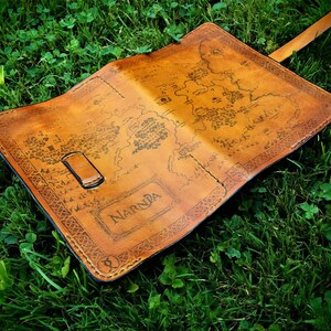 May include: A brown leather journal with a map of Narnia printed on the cover. The journal has a strap closure and is lying on a bed of green grass.