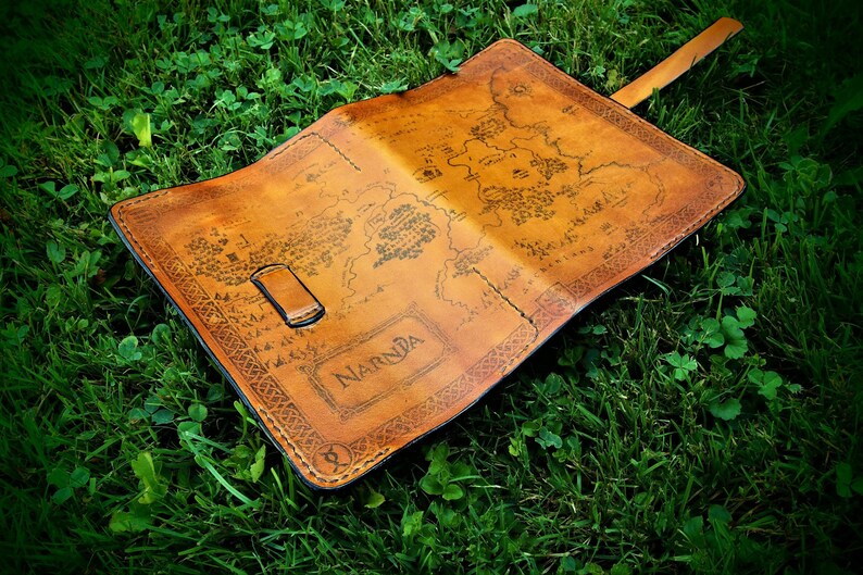 May include: A brown leather journal with a map of Narnia printed on the cover. The journal has a strap closure and is lying on a bed of green grass.