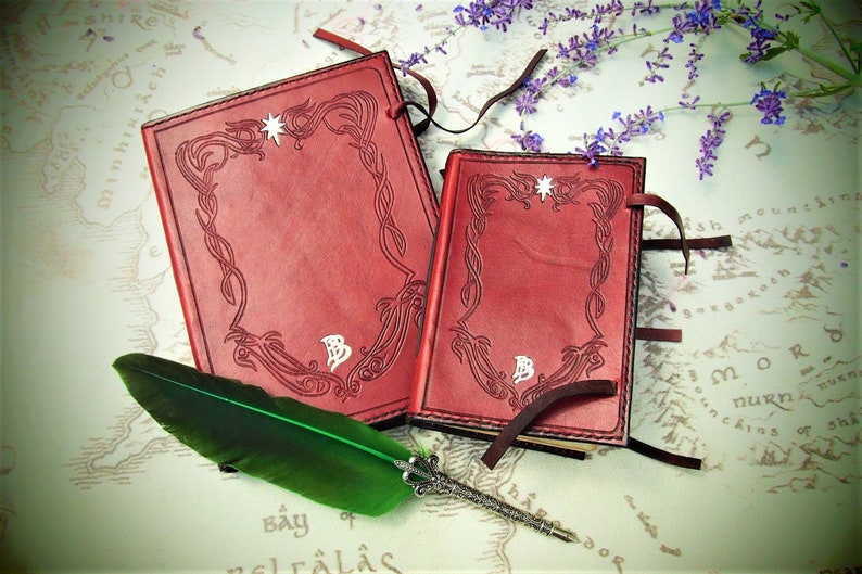 Puede incluir: Dos diarios de cuero rojo con dise&ntilde;os decorativos en relieve y acentos de estrella plateada. Los diarios tienen lazos de cuero marr&oacute;n y est&aacute;n sobre un mapa con una pluma de pluma verde.