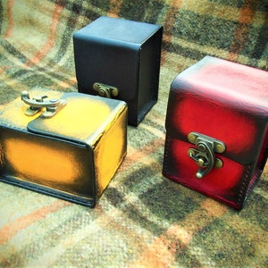 May include: Three leather boxes with different colored finishes. The boxes are closed and have metal clasps. The boxes are on a plaid fabric background.