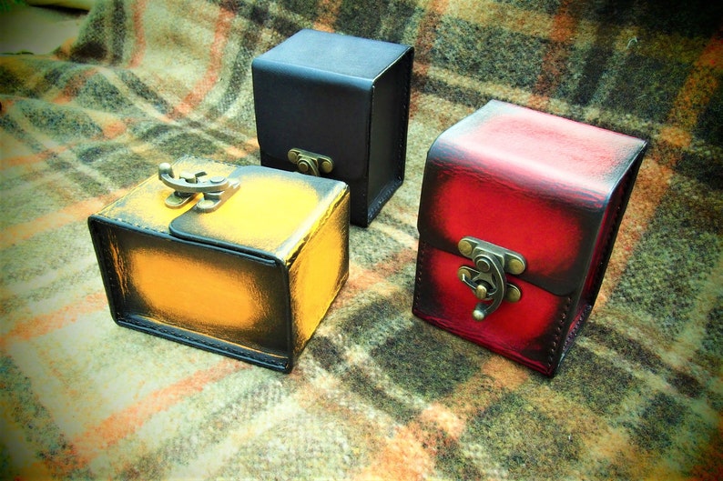 May include: Three leather boxes with different colored finishes. The boxes are closed and have metal clasps. The boxes are on a plaid fabric background.