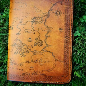 May include: A brown leather journal with a map of Middle-earth printed on the cover. The map includes details like the Shire, Rivendell, and Mordor. The journal has a Celtic knot border and a small axe symbol on the bottom right corner.