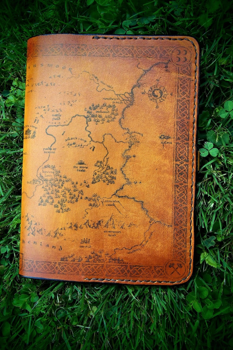 May include: A brown leather journal with a map of Middle-earth printed on the cover. The map includes details like the Shire, Rivendell, and Mordor. The journal has a Celtic knot border and a small axe symbol on the bottom right corner.