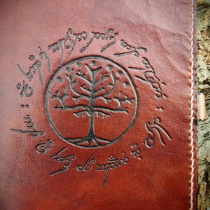 Puede incluir: Un diario de cuero marrón con un detalle de costura blanca en el borde. La portada presenta un diseño circular con un árbol y la inscripción "Ash nazg durbatulûk, ash nazg gimbatul, ash nazg thrakatulûk, agh burzum-ishi krimpatul."