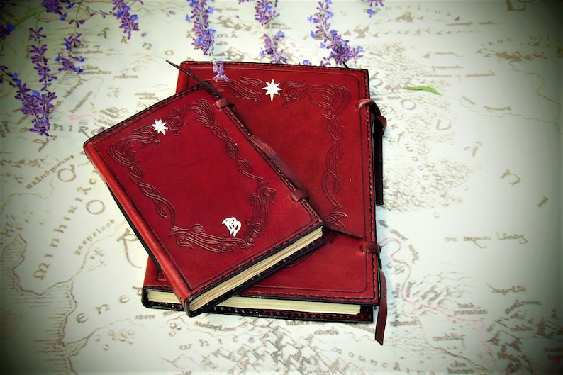 Puede incluir: Dos diarios encuadernados en cuero rojo con dise&ntilde;os decorativos en relieve y detalles de estrellas blancas. El diario m&aacute;s peque&ntilde;o tiene un s&iacute;mbolo blanco en la portada.