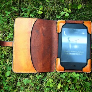 May include: A brown leather case with a flap closure for an e-reader. The e-reader is open and shows a screen with the text "Find your next favourite book".