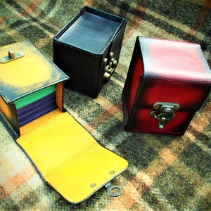 May include: Three leather boxes with different colored finishes. The box on the left is open and has a yellow interior with a green and purple stack of cards inside. The box in the center is black and the box on the right is red.