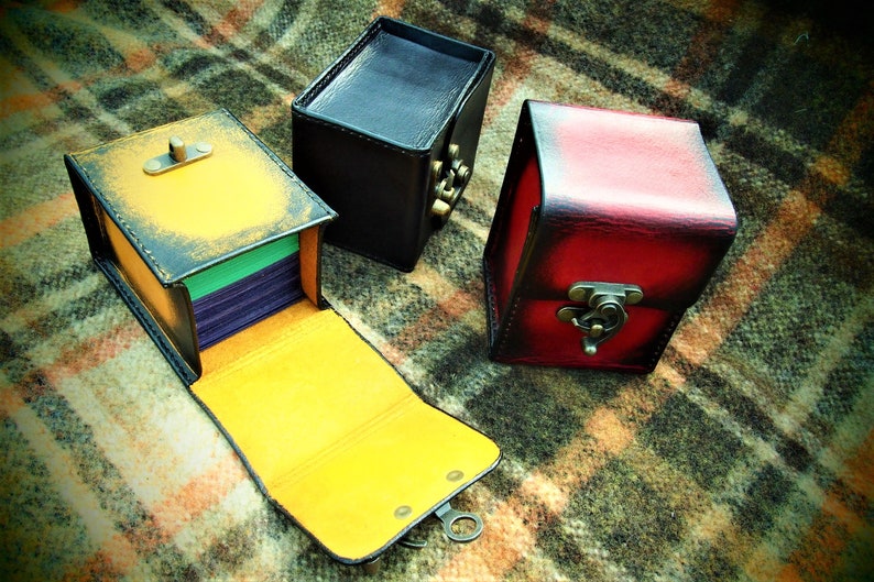 May include: Three leather boxes with different colored finishes. The box on the left is open and has a yellow interior with a green and purple stack of cards inside. The box in the center is black and the box on the right is red.