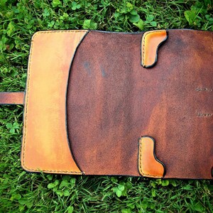 May include: A brown leather notebook cover with a flap closure and a strap. The cover has a light brown border and stitching around the edges. The cover is lying on a bed of green grass.