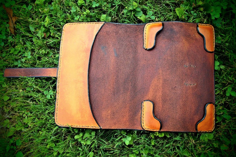 May include: A brown leather notebook cover with a flap closure and a strap. The cover has a light brown border and stitching around the edges. The cover is lying on a bed of green grass.