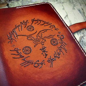 Może przedstawiać: Brązowy skórzany dziennik z białym szwem wokół krawędzi. Okładka zdobiona jest wytłoczonym wzorem smoka z rozpostartymi skrzydłami, otoczonym okręgiem tekstu w starożytnym języku. Dziennik ma brązowy skórzany pasek.