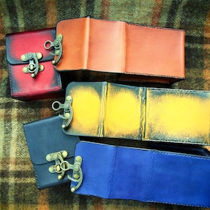 May include: Four leather boxes with different colored covers and metal clasps. The boxes are arranged in a stack, with the red box on top, followed by the orange, yellow, and blue boxes.