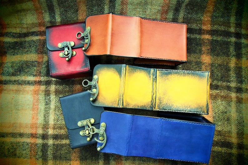May include: Four leather boxes with different colored covers and metal clasps. The boxes are arranged in a stack, with the red box on top, followed by the orange, yellow, and blue boxes.