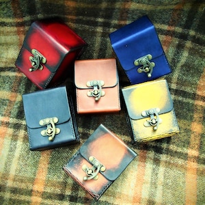 May include: Six small leather boxes with metal clasps in various colors, including red, blue, brown, yellow, and black. The boxes are arranged on a plaid fabric background.