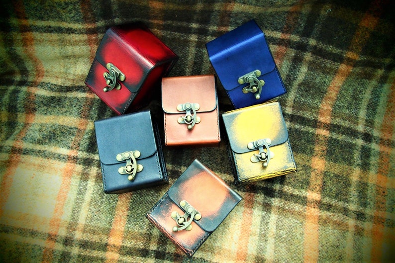 May include: Six small leather boxes with metal clasps in various colors, including red, blue, brown, yellow, and black. The boxes are arranged on a plaid fabric background.