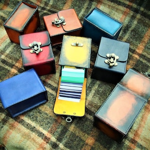 May include: A collection of nine small, leather-bound boxes with metal clasps. The boxes are in various colors, including red, blue, yellow, and brown. Some of the boxes are open, revealing a stack of cards inside.
