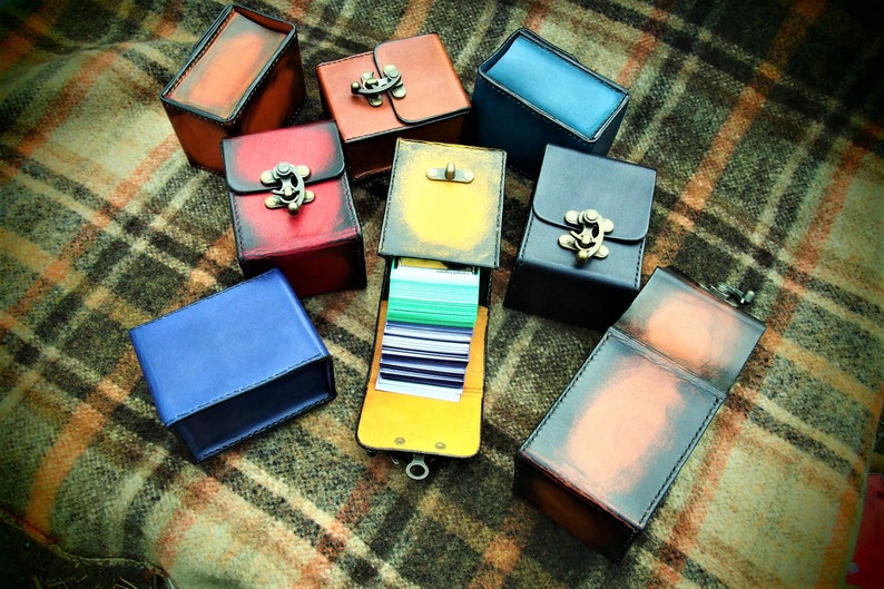 May include: A collection of nine small, leather-bound boxes with metal clasps. The boxes are in various colors, including red, blue, yellow, and brown. Some of the boxes are open, revealing a stack of cards inside.