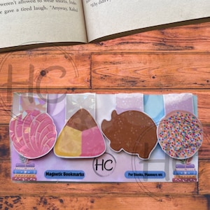 May include: A set of four magnetic bookmarks with food-themed designs. The designs include a pink concha, a candy corn, a brown pig, and a cookie with sprinkles. The text "Magnetic Bookmarks" and "For Books, Planners etc" are also visible.