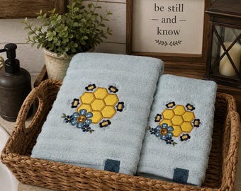 May include: Two light blue towels featuring an embroidered design of yellow honeycombs surrounded by small dark blue bees and a blue flower. The towels are folded and placed inside a rectangular woven basket with two side handles.