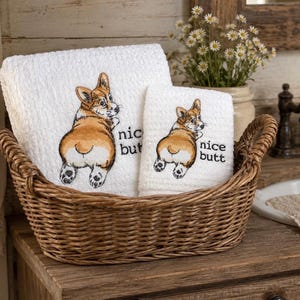May include: Two white towels featuring an embroidered illustration of a Corgi dog looking back over its shoulder with the text "nice butt" in black lettering. The towels are folded and placed inside a brown woven wicker basket.