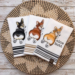 May include: Three white waffle-weave kitchen towels featuring embroidered Corgi dogs looking back over their shoulders. Each towel displays a different colored Corgi: black and white, brown and white, and tan and white. The text nice butt is embroidered on the third towel. Each towel has horizontal stripes at the bottom.