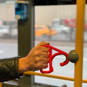 Op de afbeelding: Een rode plastic handgreep voor openbaar vervoer. De handgreep is ontworpen om mensen te helpen oppervlakken met hun handen te vermijden.