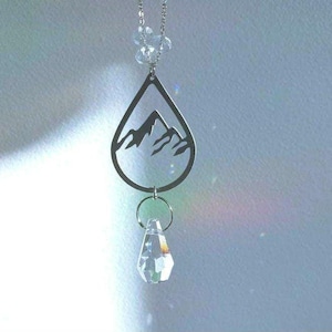 May include: A silver teardrop-shaped pendant with a mountain silhouette design hangs from a silver chain. A clear crystal dangles from the bottom of the pendant.