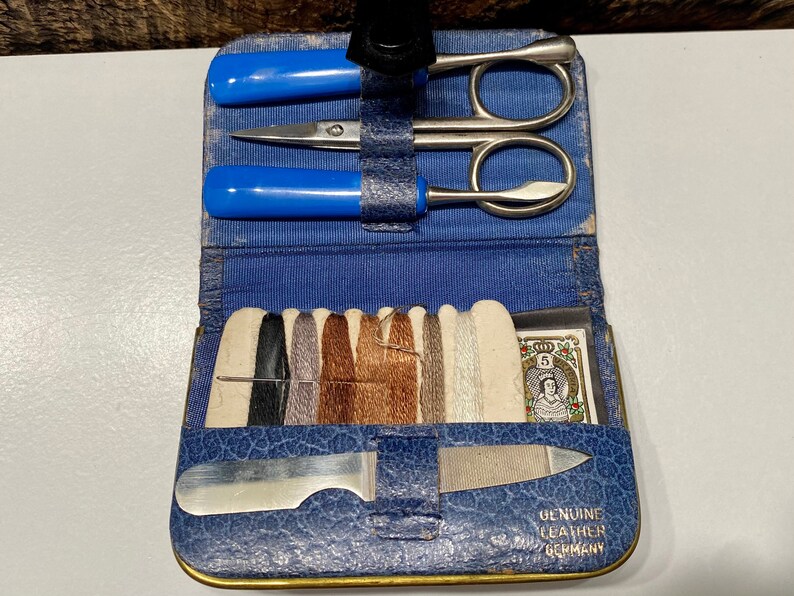 May include: A blue leather sewing kit with a variety of tools, including scissors, a needle, thread, and a nail file. The kit also includes a small card with a picture of a woman and the text "Genuine Leather Germany".
