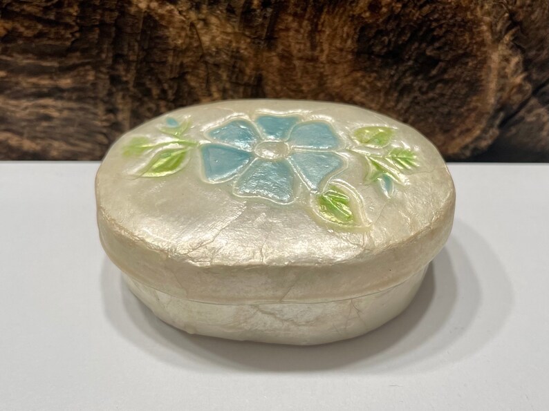 May include: A small, oval-shaped trinket box made of white paper mache with a blue flower and green leaves design.