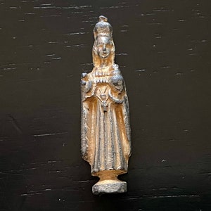 May include: A small, silver-colored lead figurine of a woman wearing a crown and a long robe. The figurine is standing on a small base.