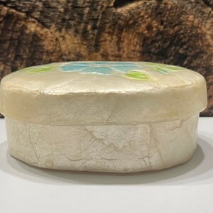 May include: A small, round, white trinket box with a floral design. The box is made of paper mache and has a textured surface. The flowers are blue, green, and white.