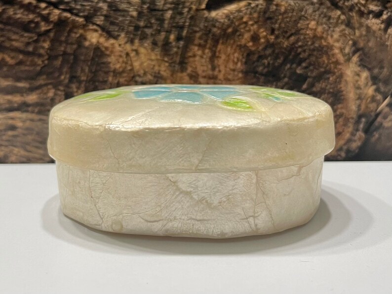 May include: A small, round, white trinket box with a floral design. The box is made of paper mache and has a textured surface. The flowers are blue, green, and white.