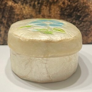 May include: A small, oval-shaped trinket box made of white mother of pearl with a floral design on the lid. The design features a blue flower with green leaves.