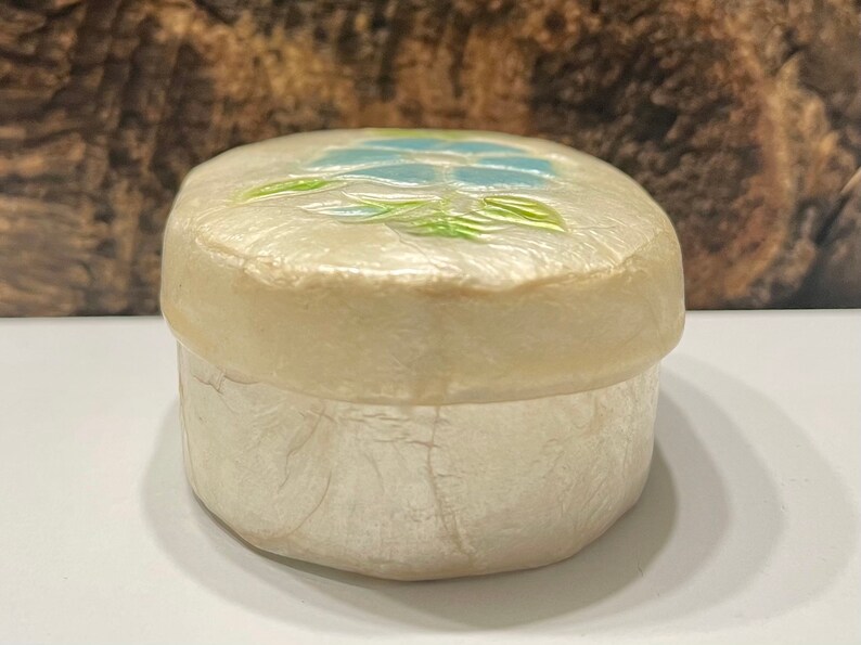 May include: A small, oval-shaped trinket box made of white mother of pearl with a floral design on the lid. The design features a blue flower with green leaves.
