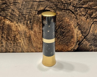 May include: A black lipstick tube with a gold top and a star pattern. The tube is on a wooden surface.