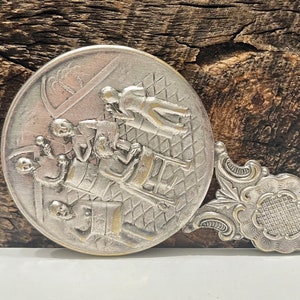 May include: A silver-toned hand mirror with a decorative handle. The mirror features a raised design of people playing a game. The handle is ornate and has a floral design.
